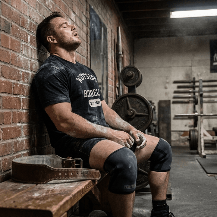 Athlete resting between sets on a gym bench wearing the black Westside Barbell EST. 1972 heavyweight vintage tee, chalk on forearms with a lifting belt and knee sleeves nearby