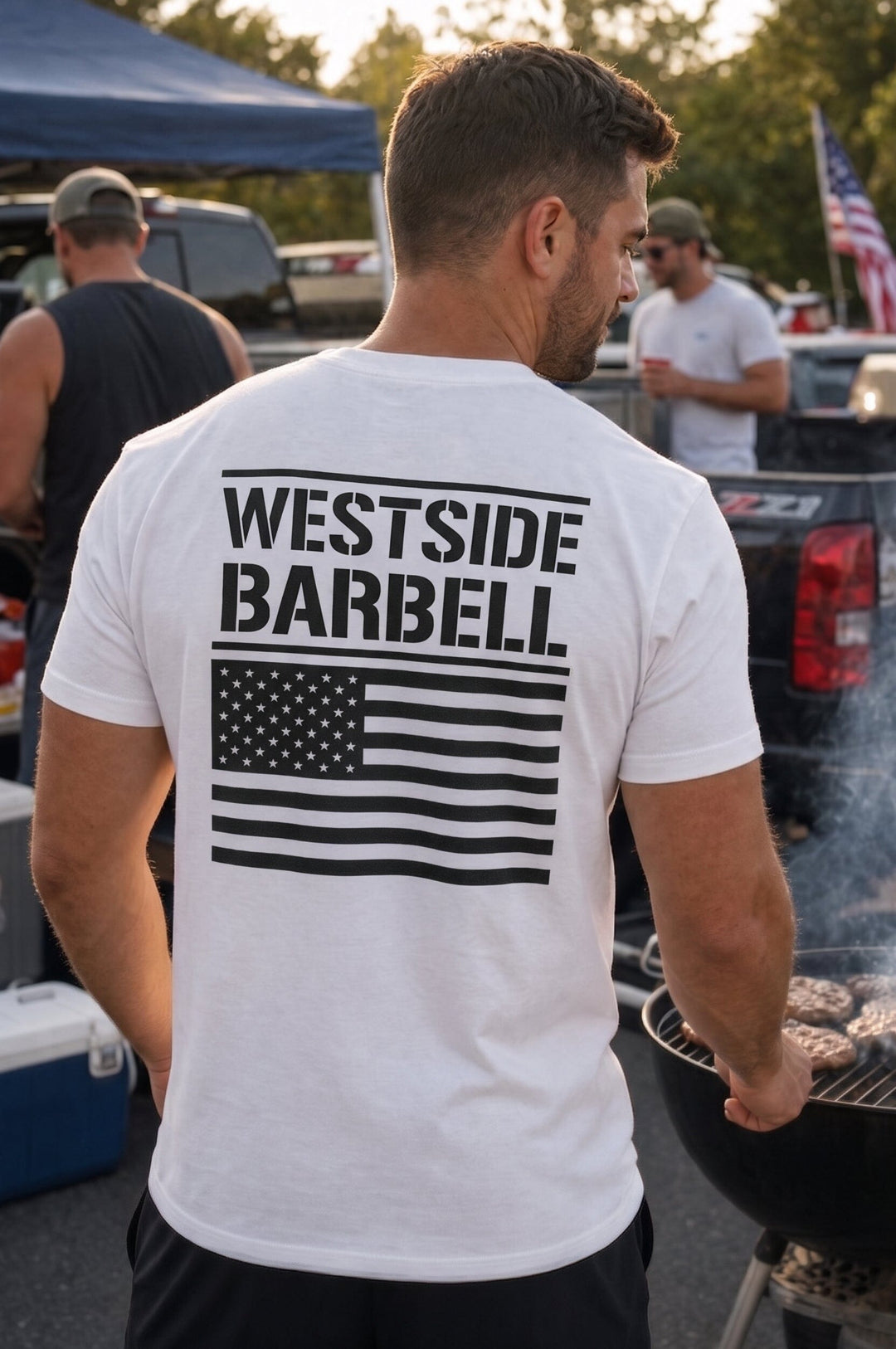 Man at an outdoor cookout seen from behind wearing the white Westside Barbell Fourth of July tee, showing the black Westside Barbell stencil text and American flag print across the back.
